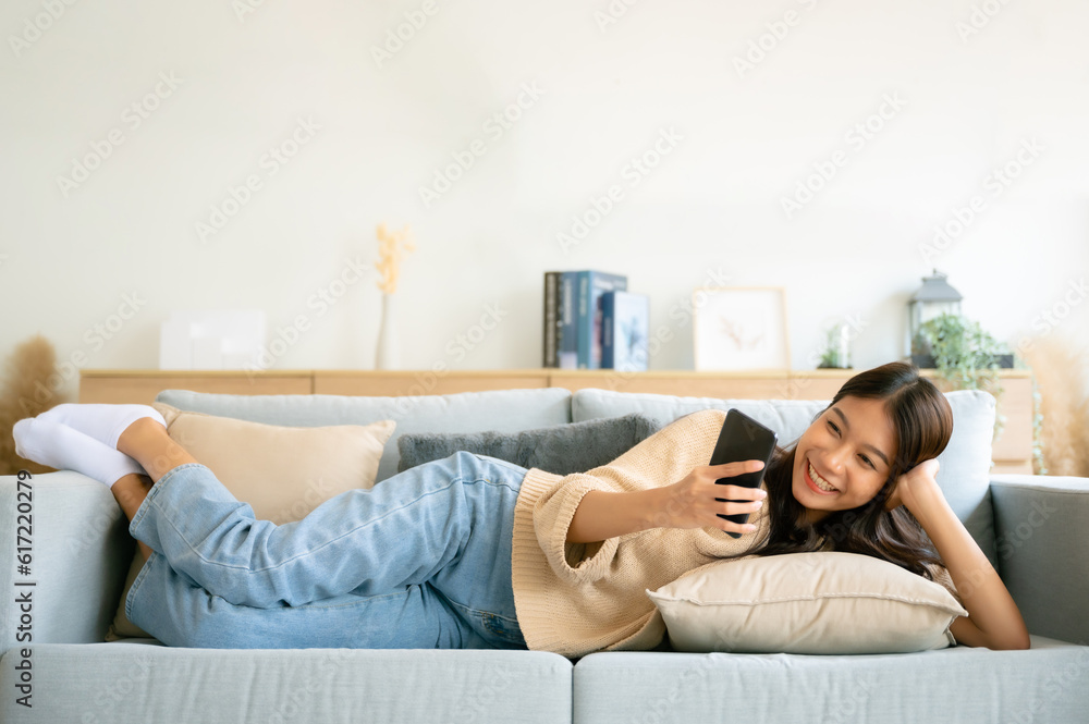 Happy young asian woman relax on comfortable couch at home texting messaging on smartphone, smiling girl use cellphone, browse wireless internet on gadget, shopping online from home Happy young asian woman relax on comfortable couch at home texting messaging on smartphone, smiling girl use cellphone, browse wireless internet on gadget, shopping online from home
