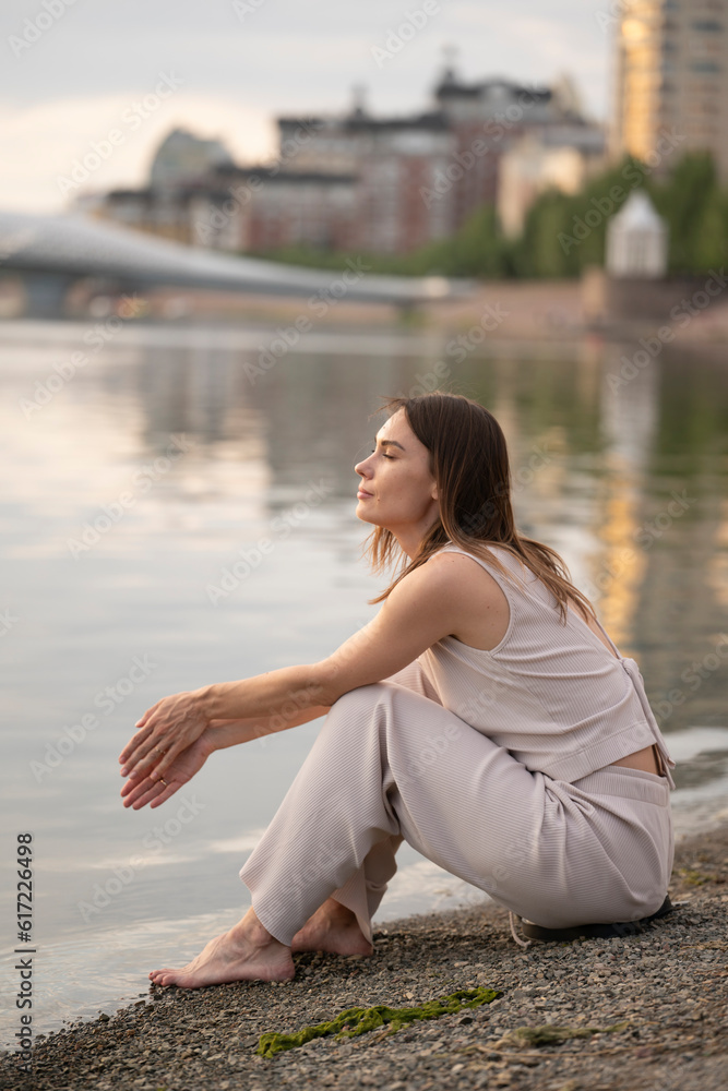 Summer urban portrait of a young brunette woman. The woman sits on the bank of the river and thinks about something.