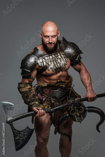 A bald bearded viking in lightweight fur-lined armor holding a large two-handed axe stands against a gray background