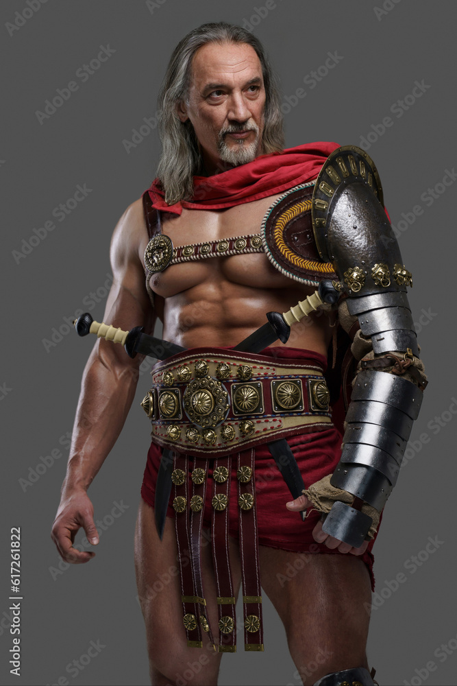 Mature and muscular gladiator with a stylish grey beard and flowing silver locks dons lightweight armor posing against grey backdrop Mature and muscular gladiator with a stylish grey beard and flowing silver locks dons lightweight armor posing against grey backdrop