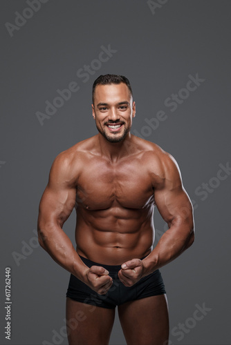 Breathtakingly handsome male model with a chiseled physique flashes a charming smile as he poses in black briefs against grey background, emanating charisma and sex appeal