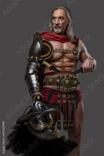 Distinguished gladiator with a stylish silver beard and luscious locks wears lightweight armor, holding gladiator helmet with feathered plume as he stands confidently against grey backdrop