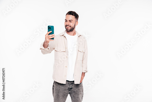 Handsome smiling stylish hipster lambersexual model. Sexy man in white T-shirt and jeans. Fashion male isolated on white background in studio. Holding smartphone, using cellphone apps, mobile phone