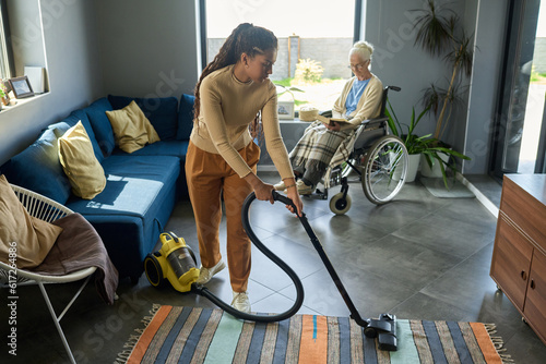 Youthful girl with vacuum cleaner helping her grandmother with disability with domestic chores while senior woman reading book by window