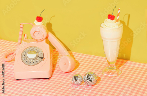 Summer creative layout with pink retro telephone with whipped cream and bright red cherry, milkshake and disco balls on pastel pink plaid and yellow background. 80s or 90s retro aesthetic girl idea. 