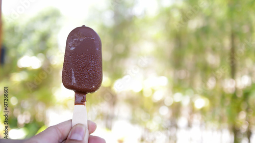 A hand holding common popular dark chocolate ice cream stick or ice bream bar with nature background copy space. Magnum ice cream.