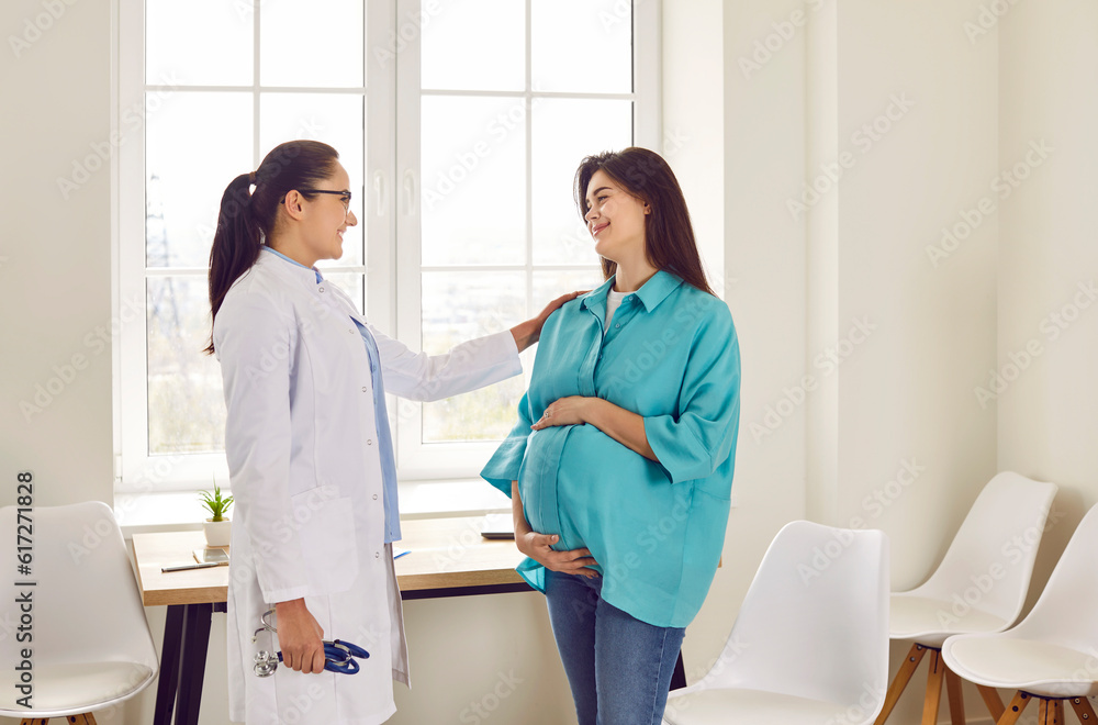 Obraz premium Doctor supporting pregnant woman. Pregnant lady visits doctor at clinic. Friendly, smiling obstetrician in white medical coat puts hand on shoulder of young pregnant girl. Pregnancy healthcare concept
