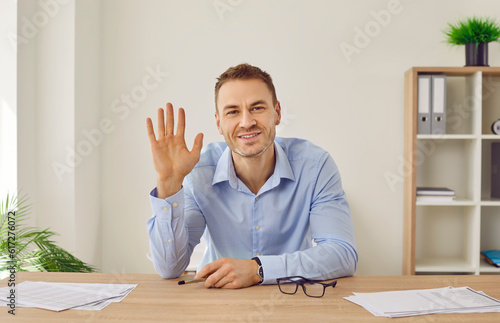 Portrait of happy man in formal blue shirt sitting at office desk in front of webcamera, making video call, starting online business meeting, looking at you, waving hand, saying hello and smiling