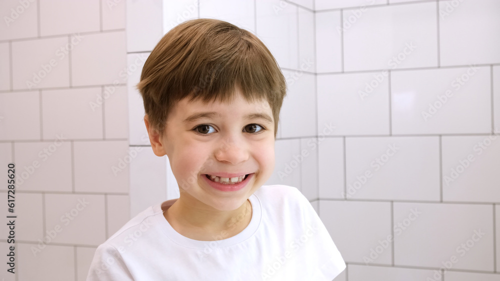 Happy child looks at the camera and smiles. Cute caucasian 6 year old boy in white t-shirt. Close-up portrait. Concept of happiness and good mood. Cheerful kid. Pretty face. Home bathroom background Happy child looks at the camera and smiles. Cute caucasian 6 year old boy in white t-shirt. Close-up portrait. Concept of happiness and good mood. Cheerful kid. Pretty face. Home bathroom background