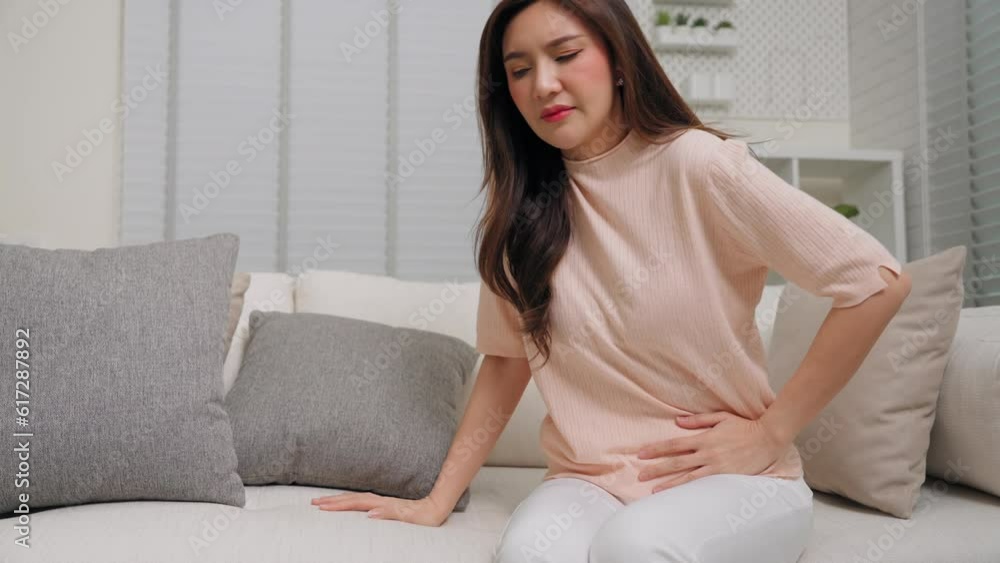 Asian female abdominal pain and nausea sitting on sofa in living room at home. Asian woman having menstruation feeling belly pain and shoulder slouching at home. Health care concept Asian female abdominal pain and nausea sitting on sofa in living room at home. Asian woman having menstruation feeling belly pain and shoulder slouching at home. Health care concept