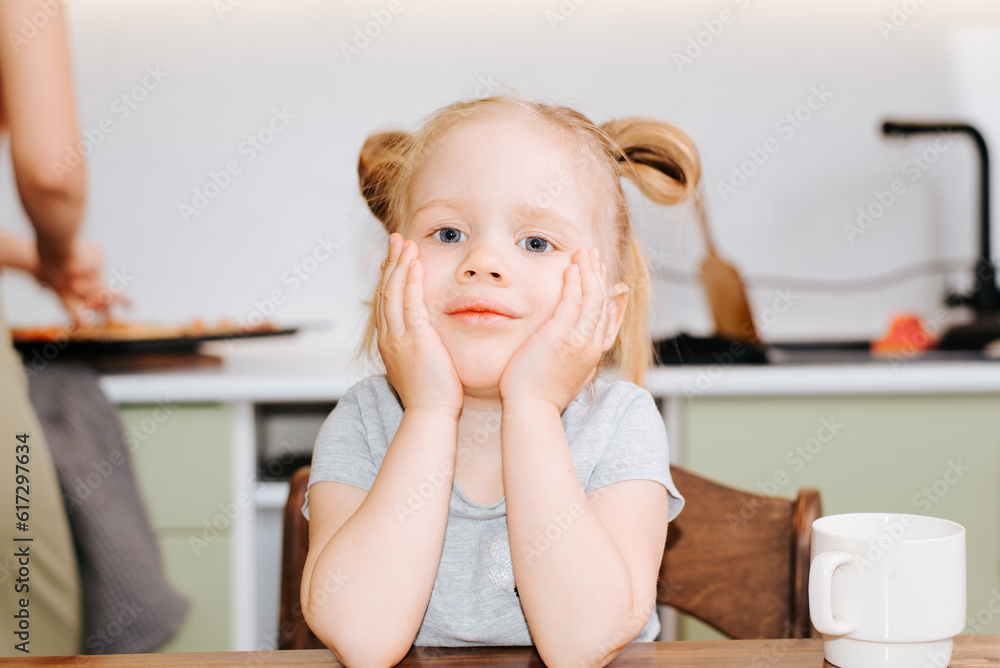 Fototapeta premium Portrait sad caucasian girl sitting at the table in the kitchen, upset hungry child waiting for food indoors