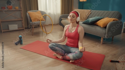 A young woman sits crosslegged in a lotus position with her eyes closed. A redhaired woman in a pink tracksuit, headband, and white wireless headphones practices yoga at home. Sports concept.