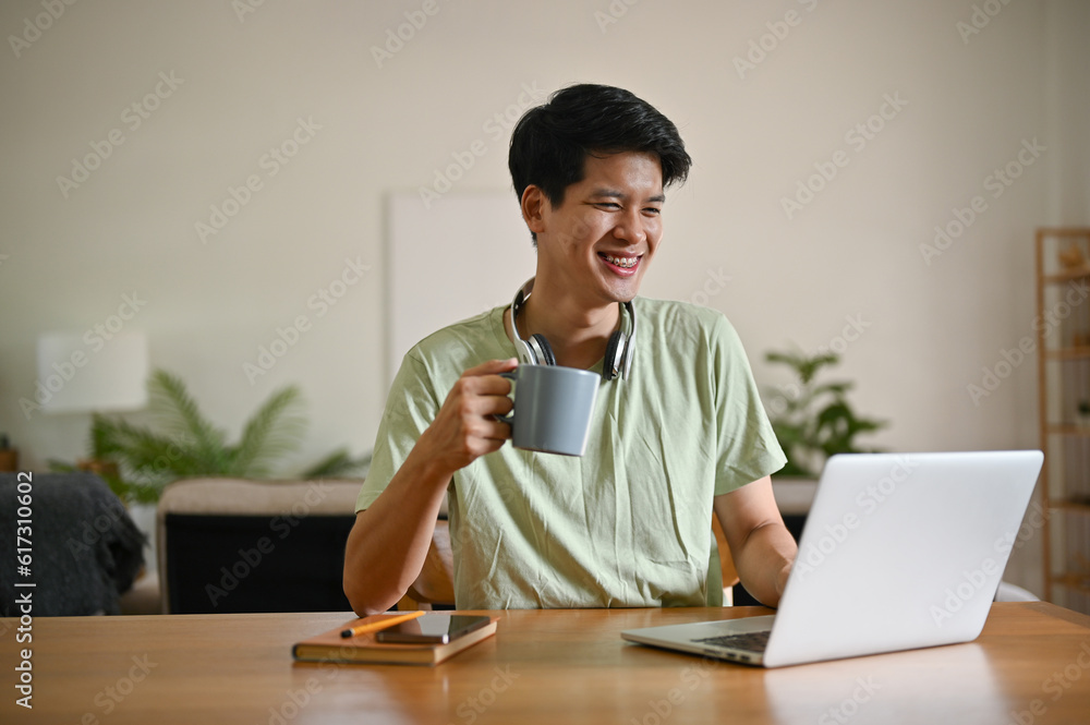 A happy young Asian man is sipping coffee while looking down at his laptop screen at a table