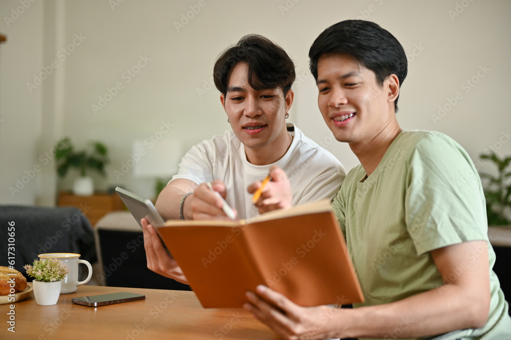 A smart Asian male college student is tutoring to his friend while studying at home together. A smart Asian male college student is tutoring to his friend while studying at home together.