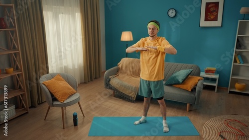 Sports at home. A man in sportswear and with a green headband is warming up in the living room. The man holds his arms bent at the elbows in front of him at chest level. Home fitness concept.