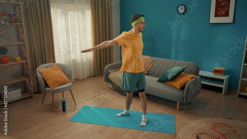 Sports at home. A man in sportswear and with a green headband is warming up in the living room. The man took his straight arms back, connecting the shoulder blades and making turns of the torso. Home