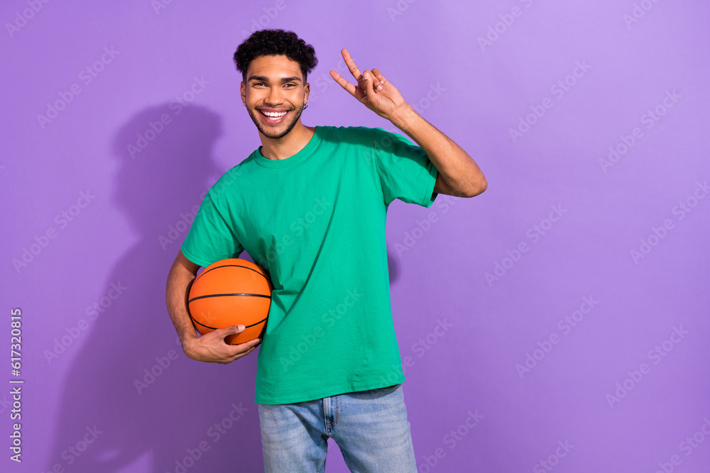 Photo of young handsome guy wear green t-shirt show v-sign hold ball professional basketball player isolated on violet color background