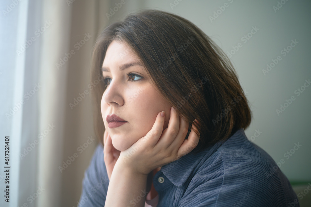 Sad plump or overweight young girl looks out window portrait. Problems of loneliness and mental health plus size people in society.