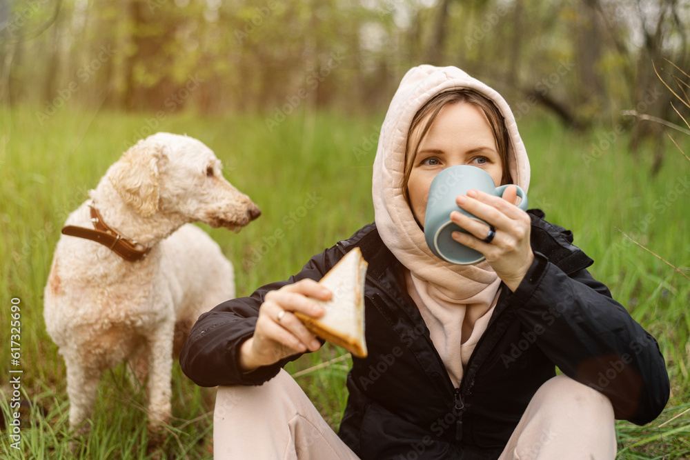 Woman eating sandwich at picnic in green forest. Her dog is nearby. Woman eating sandwich at picnic in green forest. Her dog is nearby.