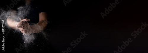 A man claps his hands with scattering flour against a dark background. Culinary theme. Banner. Place for your text.