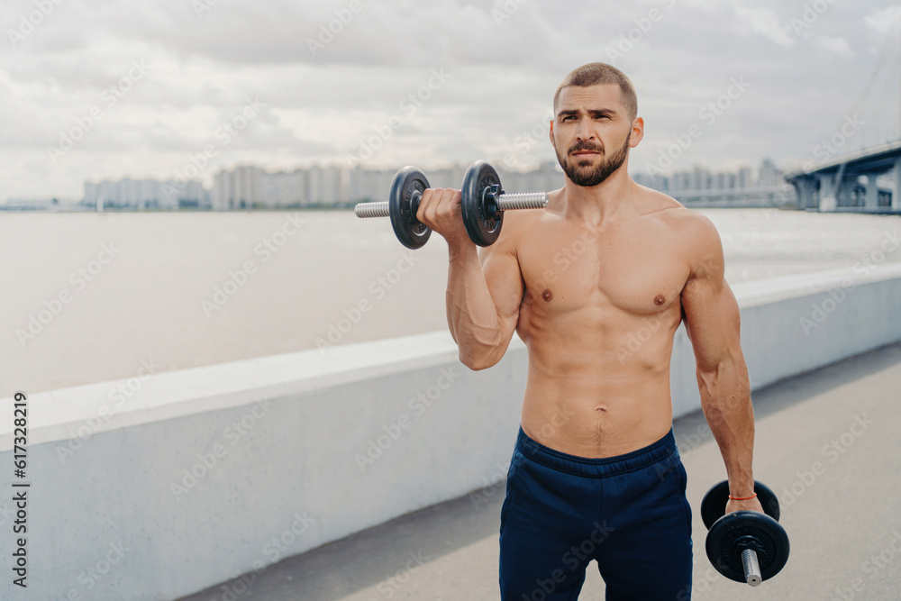 Handsome bearded man pumps muscles, exercises with barbells, leads sporty lifestyle, has strong body. Outdoor near river, bare-chested bodybuilder lifts heavy dumbbells. Handsome bearded man pumps muscles, exercises with barbells, leads sporty lifestyle, has strong body. Outdoor near river, bare-chested bodybuilder lifts heavy dumbbells.