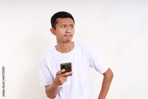 Confused asian funny man holding cell phone while looking sideways. Isolated on white background