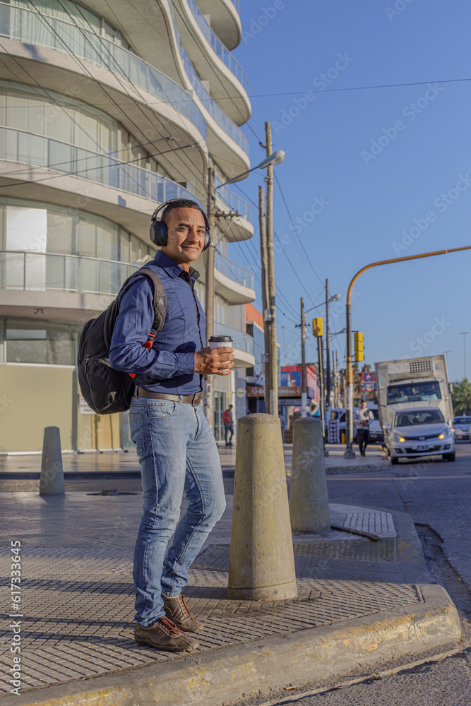 Young latin man with headphones and casual clothing crossing the street in a city.