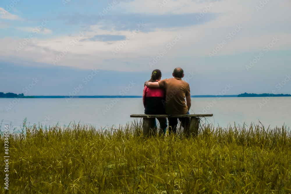 Silhouette of a couple hugging. The pair are sitting on a wooden bench on the lake background. Changed color. Copy space.