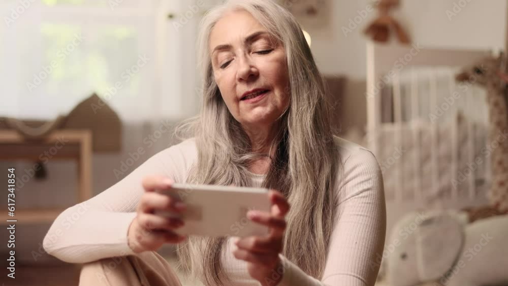 Portrait of excited senior grey haired woman gamer sitting on floor and playing in online video games on smartphone holding in horizontal landscape mode at cozy living room at home