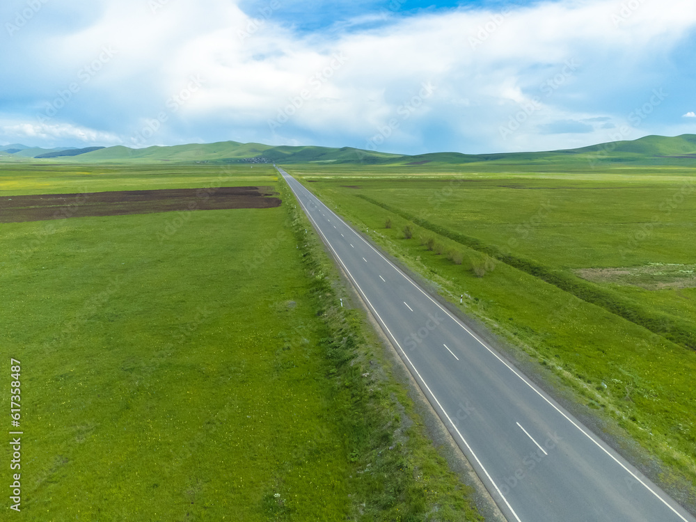 Drone view of an empty paved road in green meadows, mountains on a sunny day. Beautiful landscape with a roadway, sunlight, trees, green grass, clouds. Armenia