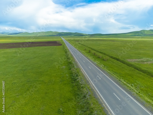 Drone view of an empty paved road in green meadows, mountains on a sunny day. Beautiful landscape with a roadway, sunlight, trees, green grass, clouds. Armenia © yaroslav1986