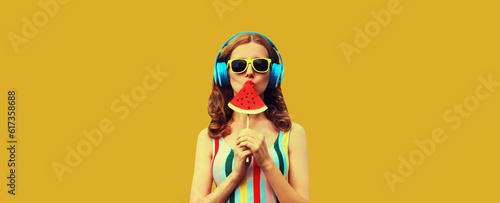 Summer portrait of stylish woman in headphones listening to music blowing her lips with juicy lollipop or ice cream shaped slice watermelon on yellow background