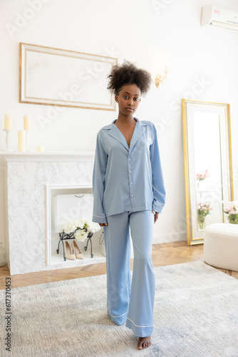 Young african woman in blue pajamas walking on carpet in bedroom.