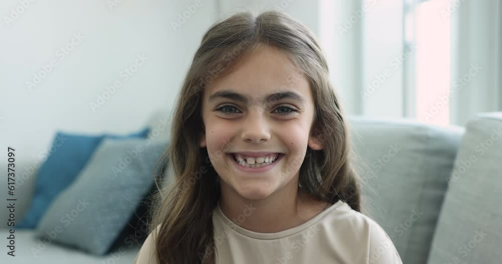 Close up head shot cute little girl sit on sofa smile look pose for camera. Video call, virtual meeting event participant or blogger, dental clinic services for children, alpha generation kid portrait Close up head shot cute little girl sit on sofa smile look pose for camera. Video call, virtual meeting event participant or blogger, dental clinic services for children, alpha generation kid portrait