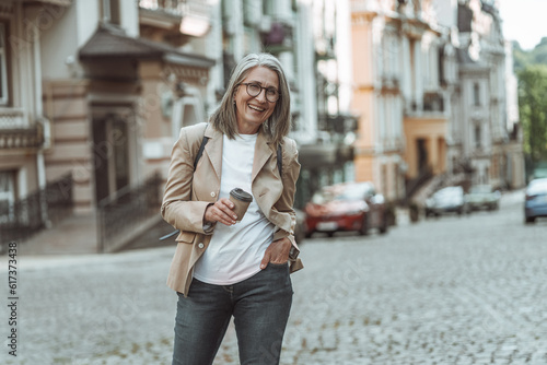 Happy senior woman captured on street of old town in Europe, holding coffee in paper cup. Smiling mature lady, with her silver hair, gracefully walks along charming streets.  © Svyatoslav Lypynskyy