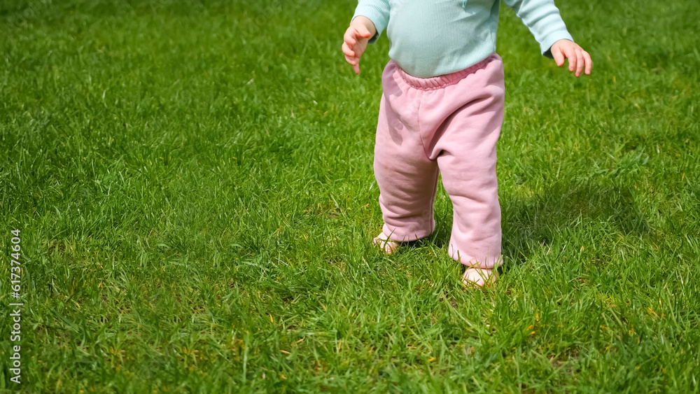 Toddler girl learns to walk independently barefoot on meadow green grass illuminated by sunlight. Child does confident steps in park Toddler girl learns to walk independently barefoot on meadow green grass illuminated by sunlight. Child does confident steps in park
