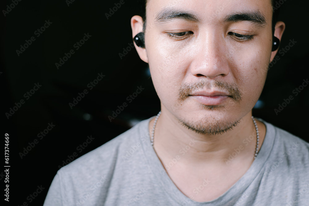 Obraz premium Close-up shot of a partial face of a young Asian man wearing a grey t-shirt listening to music on his wireless earphones. Shot from front of him. Isolated black background.