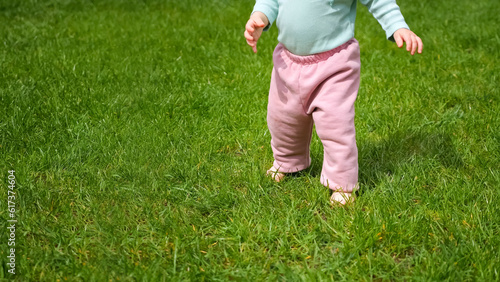 Toddler girl learns to walk independently barefoot on meadow green grass illuminated by sunlight. Child does confident steps in park © lenblr