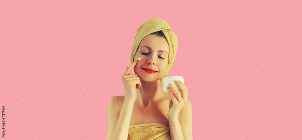 Portrait of happy smiling young caucasian woman applying face cream and drying wet hair in wrapped bath towel on pink background Portrait of happy smiling young caucasian woman applying face cream and drying wet hair in wrapped bath towel on pink background