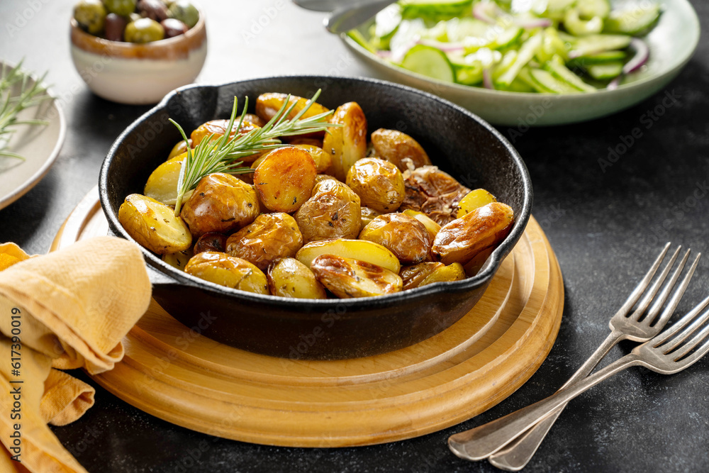 Roasted new potato with rosemary in frying pan, served with olives and fresh salad, dark background. 