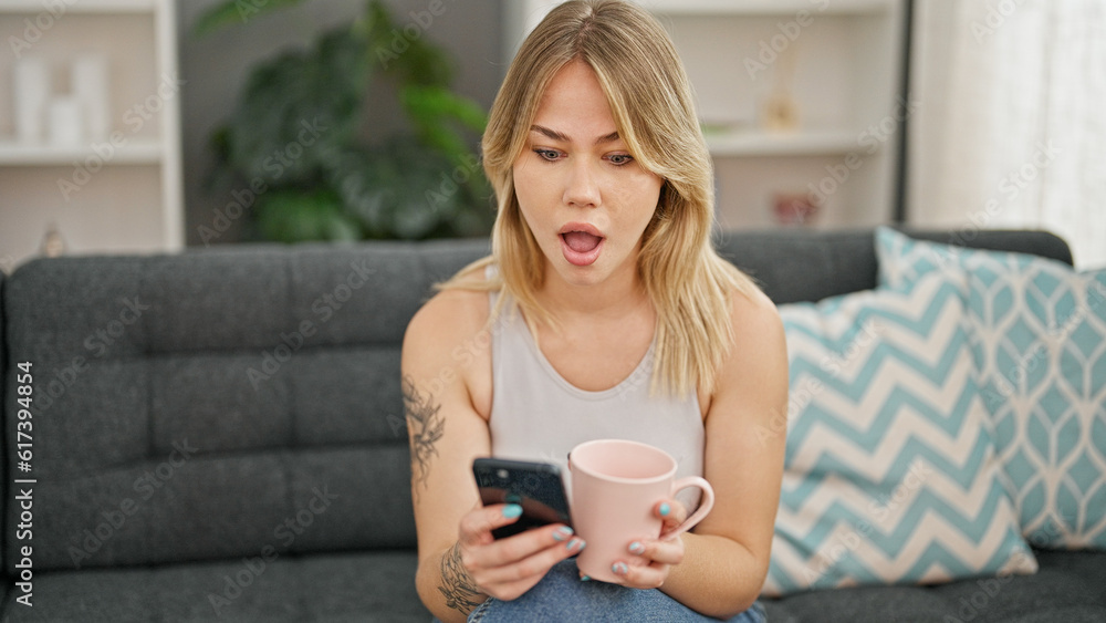 Young blonde woman using smartphone drinking coffee with surprise expression at home Young blonde woman using smartphone drinking coffee with surprise expression at home