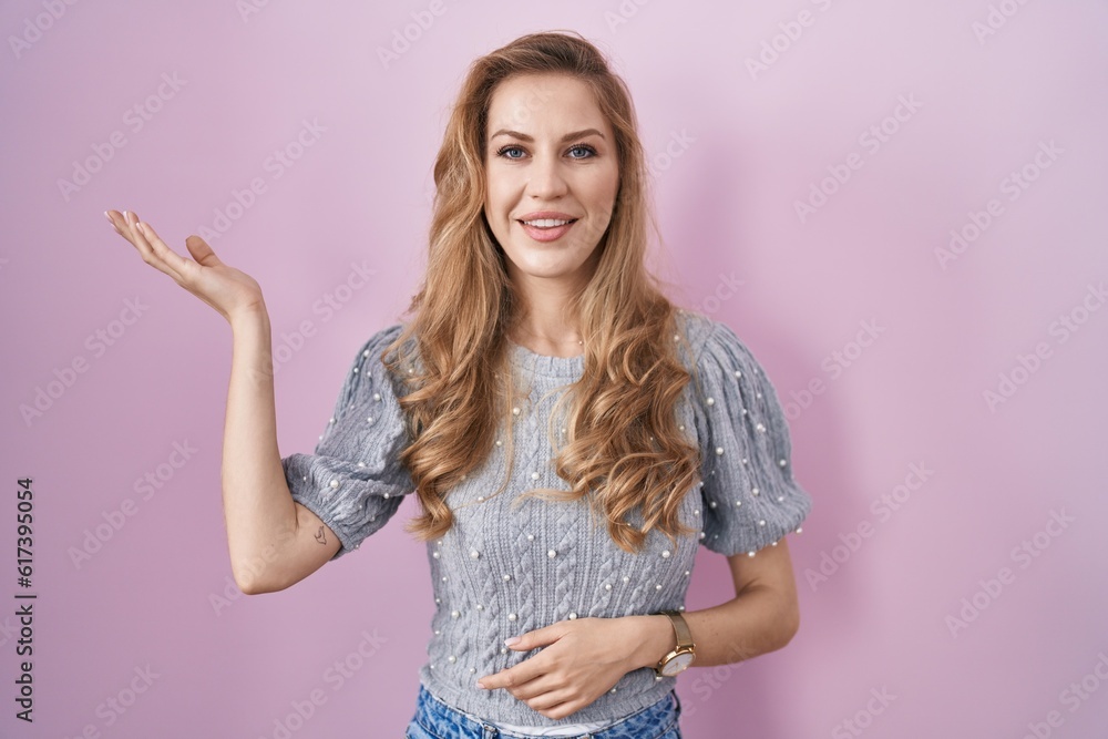 Beautiful blonde woman standing over pink background smiling cheerful presenting and pointing with palm of hand looking at the camera. Beautiful blonde woman standing over pink background smiling cheerful presenting and pointing with palm of hand looking at the camera.