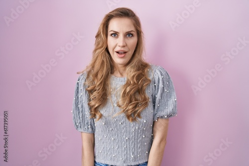 Beautiful blonde woman standing over pink background afraid and shocked with surprise and amazed expression, fear and excited face.