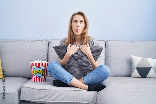 Beautiful blonde woman watching tv sitting on the sofa making fish face with mouth and squinting eyes, crazy and comical.