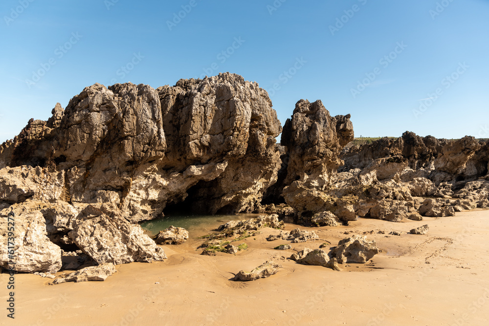 Panoramic view of a cave formed on the beach by large rocks with a small lake of fresh sea water and golden sand on the coast of Asturias on a sunny day.