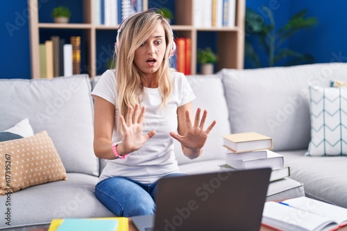 Young blonde woman studying using computer laptop at home afraid and terrified with fear expression stop gesture with hands, shouting in shock. panic concept.