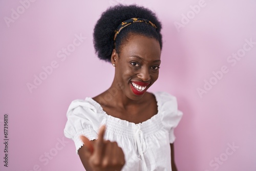 African woman with curly hair standing over pink background beckoning come here gesture with hand inviting welcoming happy and smiling