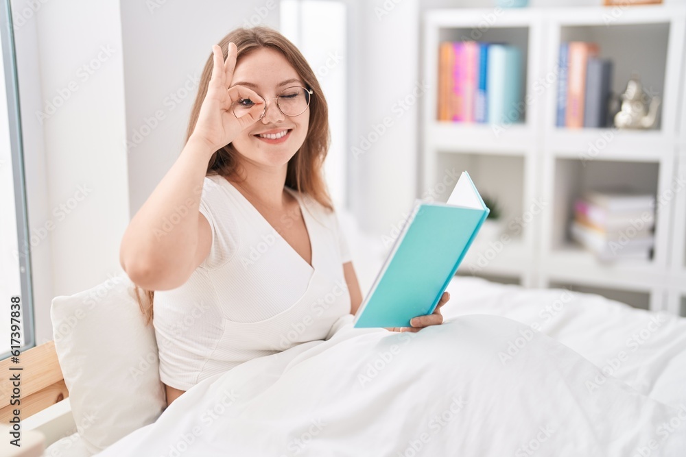 Young caucasian woman sitting on the bed at home reading a book smiling happy doing ok sign with hand on eye looking through fingers Young caucasian woman sitting on the bed at home reading a book smiling happy doing ok sign with hand on eye looking through fingers
