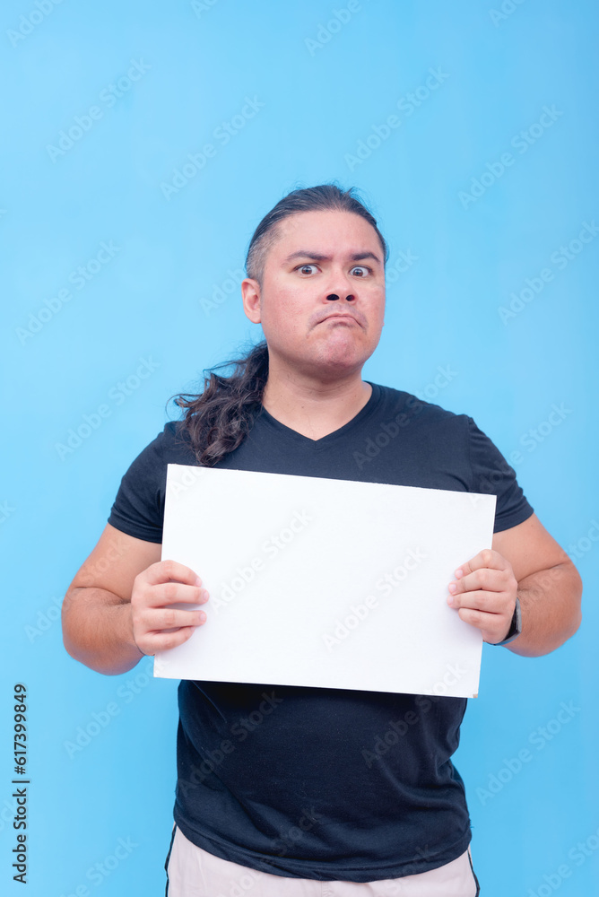 A biracial male with undercut angrily holding a blank placard with lips pursed head tilted. Isolated on a blue background. A biracial male with undercut angrily holding a blank placard with lips pursed head tilted. Isolated on a blue background.