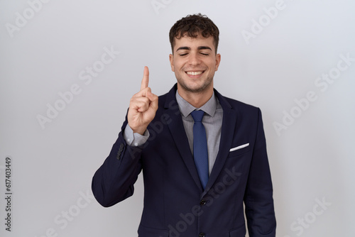 Young hispanic business man wearing suit and tie showing and pointing up with finger number one while smiling confident and happy.